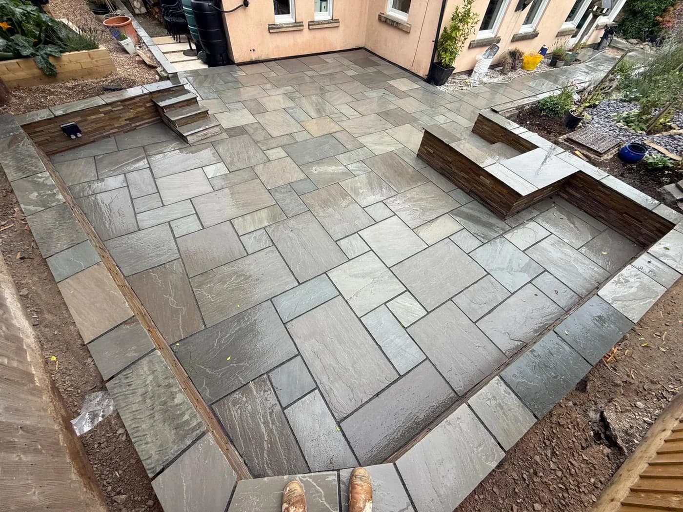 Large grey Indian sandstone patio with stone clad raised beds, built in seating and paved steps up to the house