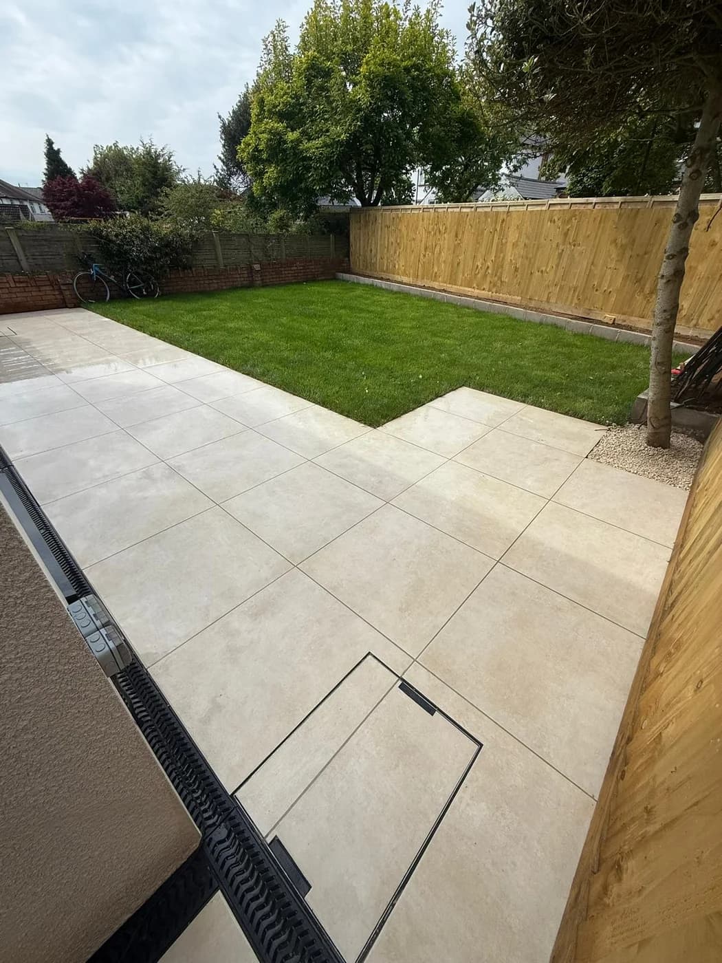 Cream large format porcelain paving with a channel drain, new turf lawn and kerb edging against a new timber fence