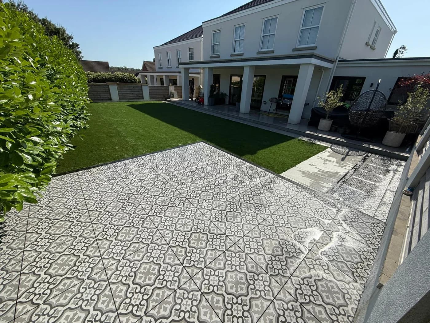 Patterned monochrome porcelain patio tiles laid beside an artificial grass lawn in the rear garden of a large modern house