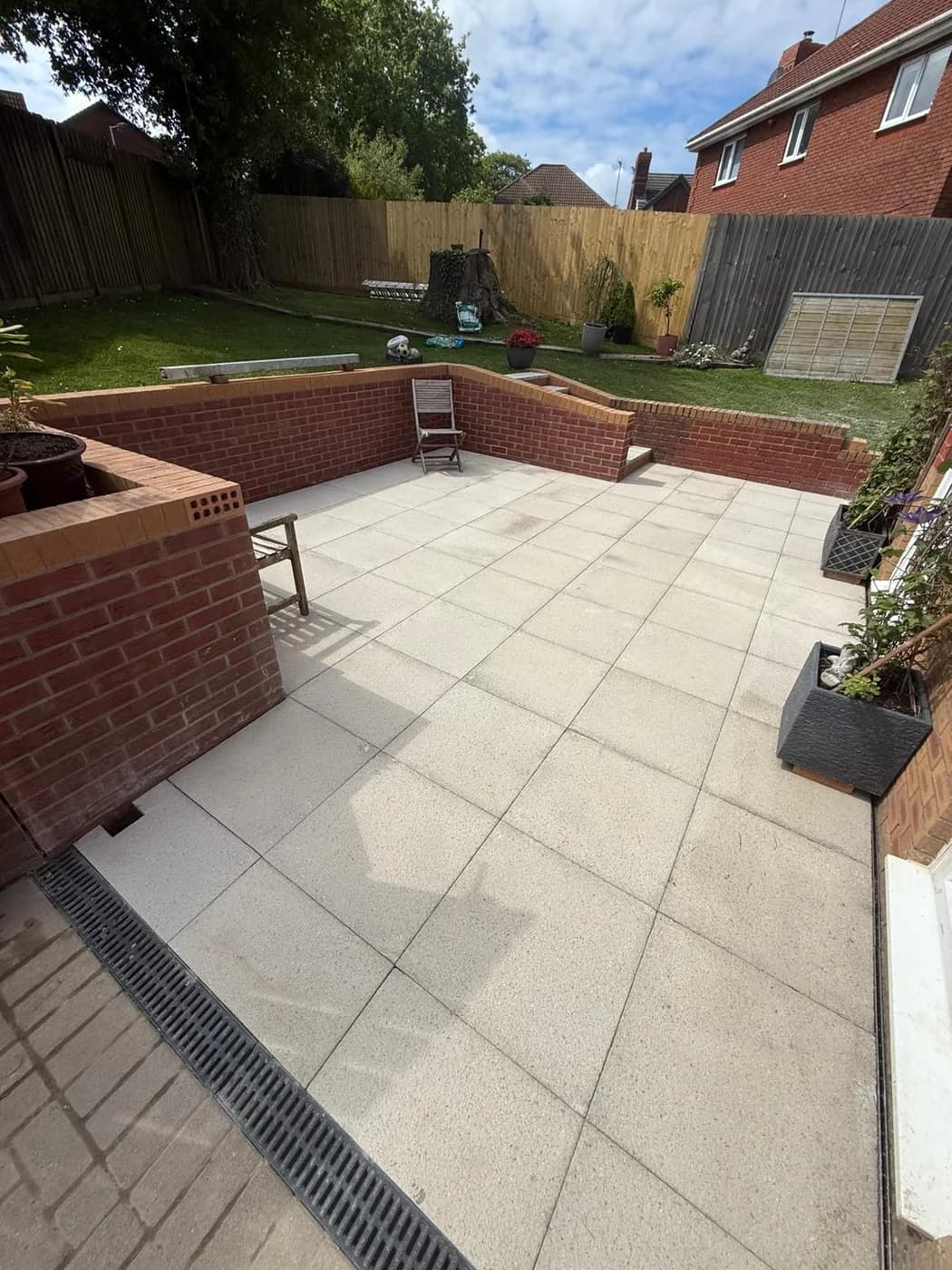 Sunken patio in light granite paving enclosed by red brick retaining walls holding back a raised lawn, with steps up to the garden